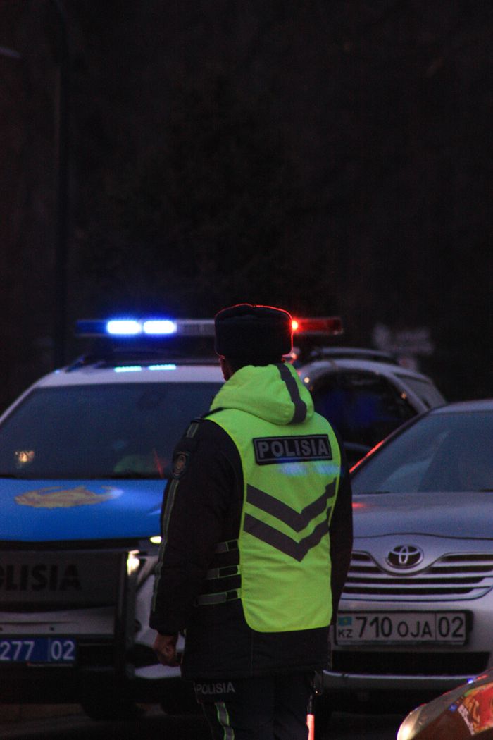Police officer in urban Almaty at night, standing by patrol cars on city street.