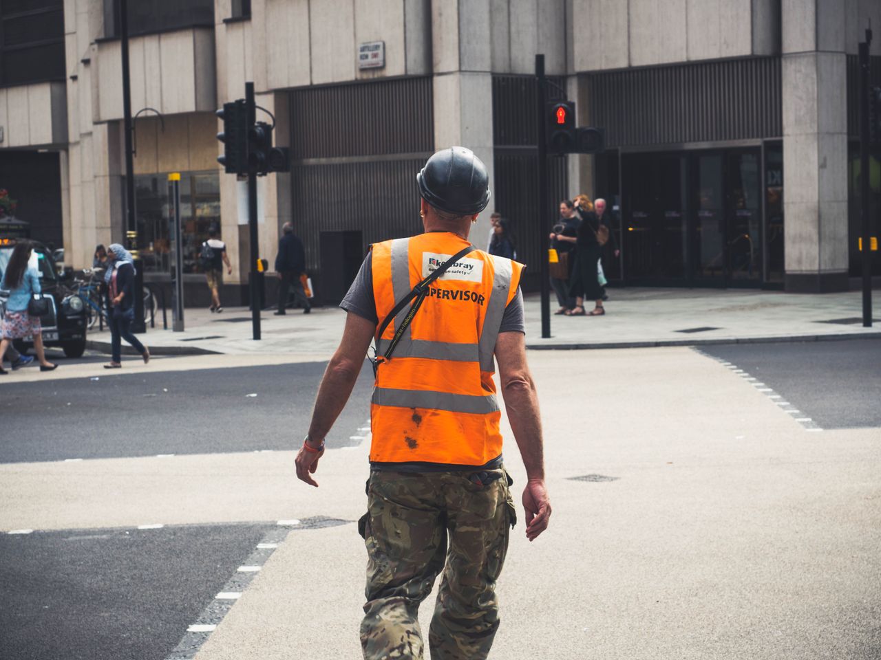 Back view of unrecognizable young male constructor in protective helmet and vest walking on asphalt road in modern city district